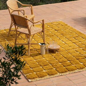 Large handmade Moroccan high-pile shag rug in saturated Thai curry mustard-gold, displayed vertically against a white brick wall. Surface features a tone-on-tone square grid pattern formed through pile variation — raised mustard squares separated by recessed ivory cream channel lines across the entire field. Dense, plush, curly wool pile texture throughout. Knotted braided ivory fringe along the top hem edge and long hand-tied ivory fringe along the bottom edge.