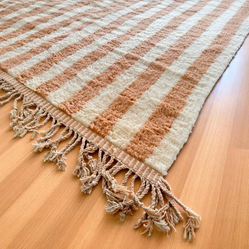 Elegant handwoven Moroccan rug with vertical stripes in Cameo Rose and soft white. Perfect for boho, coastal, minimalist, or Japandi-inspired spaces. Made from natural wool, this artisan piece blends texture and comfort for your living room, bedroom, or entryway. A modern pastel rug with warm earthy tones and timeless Amazigh character. Custom sizes available.