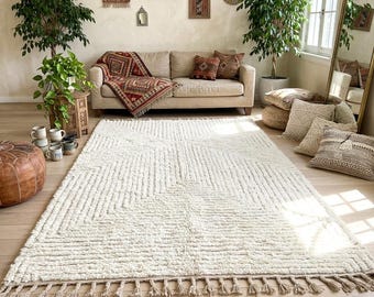 Peut inclure: Un grand tapis blanc cassé avec un motif géométrique et des franges. Le tapis est au centre d'un salon, complété par un canapé beige, des coussins décoratifs et des plantes en pot. La pièce a une esthétique bohème.