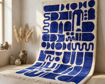 Può includere: Un tappeto rettangolare caratterizzato da una base color crema con un audace motivo geometrico blu intenso composto da varie forme, tra cui archi, cerchi e linee ondulate, rifinito con una corta frangia lungo il bordo inferiore.