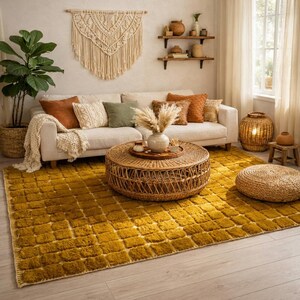 Large handmade Moroccan high-pile shag rug in saturated Thai curry mustard-gold, displayed vertically against a white brick wall. Surface features a tone-on-tone square grid pattern formed through pile variation — raised mustard squares separated by recessed ivory cream channel lines across the entire field. Dense, plush, curly wool pile texture throughout. Knotted braided ivory fringe along the top hem edge and long hand-tied ivory fringe along the bottom edge.
