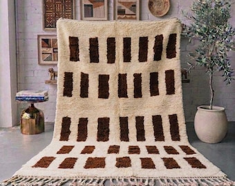 May include: A rectangular area rug featuring a cream-colored base with a repeating pattern of dark brown vertical rectangular blocks. The rug has a thick, shaggy pile texture and is finished with a fringe of cream-colored tassels along the bottom edge.