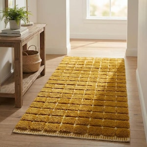 Large handmade Moroccan high-pile shag rug in saturated Thai curry mustard-gold, displayed vertically against a white brick wall. Surface features a tone-on-tone square grid pattern formed through pile variation — raised mustard squares separated by recessed ivory cream channel lines across the entire field. Dense, plush, curly wool pile texture throughout. Knotted braided ivory fringe along the top hem edge and long hand-tied ivory fringe along the bottom edge.