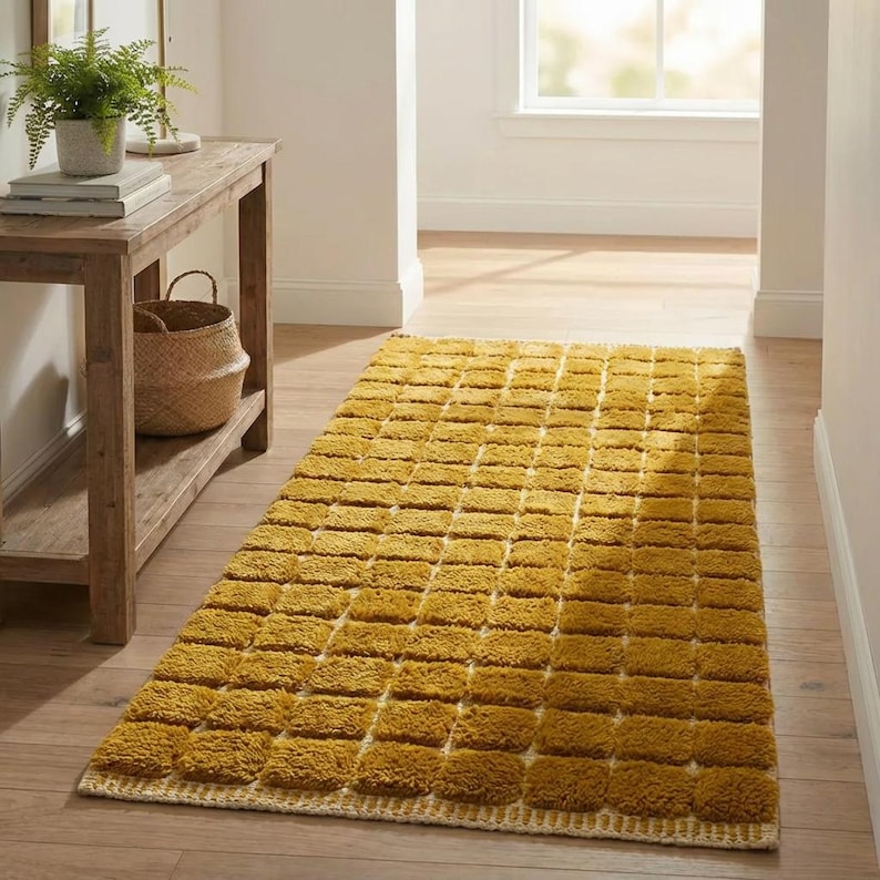 Large handmade Moroccan high-pile shag rug in saturated Thai curry mustard-gold, displayed vertically against a white brick wall. Surface features a tone-on-tone square grid pattern formed through pile variation — raised mustard squares separated by recessed ivory cream channel lines across the entire field. Dense, plush, curly wool pile texture throughout. Knotted braided ivory fringe along the top hem edge and long hand-tied ivory fringe along the bottom edge.