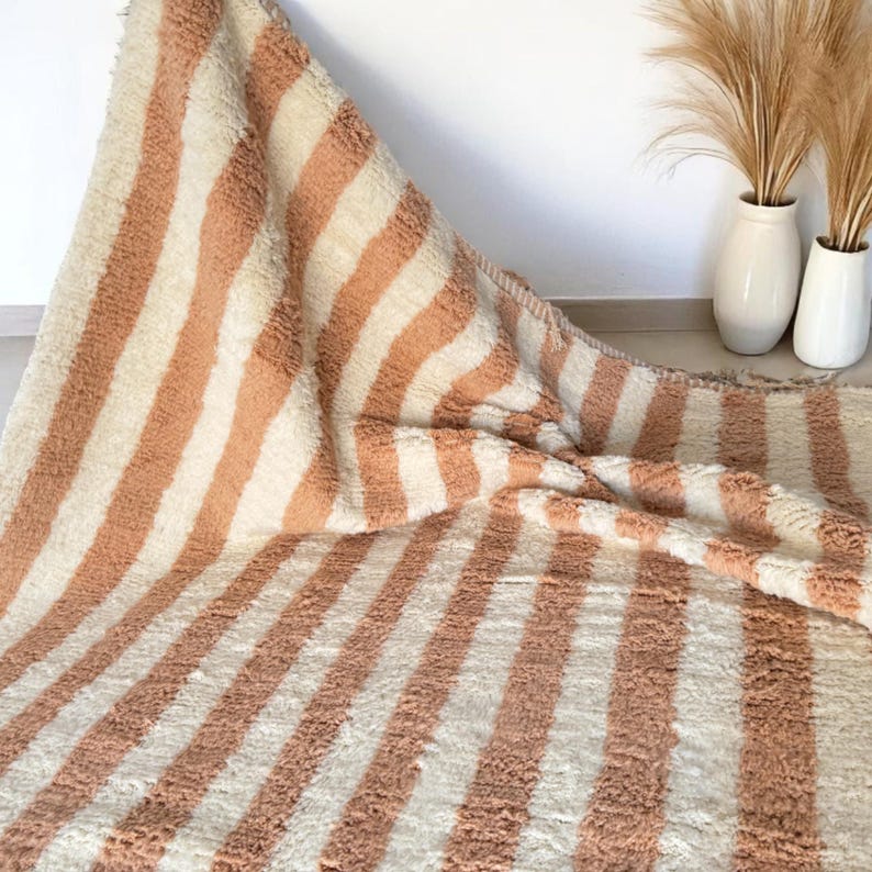 Elegant handwoven Moroccan rug with vertical stripes in Cameo Rose and soft white. Perfect for boho, coastal, minimalist, or Japandi-inspired spaces. Made from natural wool, this artisan piece blends texture and comfort for your living room, bedroom, or entryway. A modern pastel rug with warm earthy tones and timeless Amazigh character. Custom sizes available.