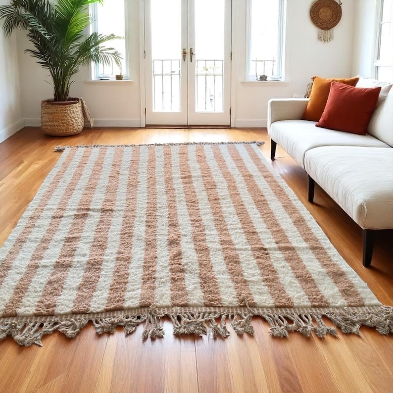 Elegant handwoven Moroccan rug with vertical stripes in Cameo Rose and soft white. Perfect for boho, coastal, minimalist, or Japandi-inspired spaces. Made from natural wool, this artisan piece blends texture and comfort for your living room, bedroom, or entryway. A modern pastel rug with warm earthy tones and timeless Amazigh character. Custom sizes available.
