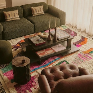 A vibrant, high-pile handmade Moroccan wool rug displayed in a bright industrial studio against a white brick wall. The rug features an eclectic, abstract tribal design with bold splashes of neon pink, teal green, orange, royal blue, and charcoal grey on a natural cream base. The patterns include geometric shapes, abstract figures, and spontaneous color blocking. The rug showcases a thick, plush shaggy texture and authentic hand-knotted cream tassels at the bottom.