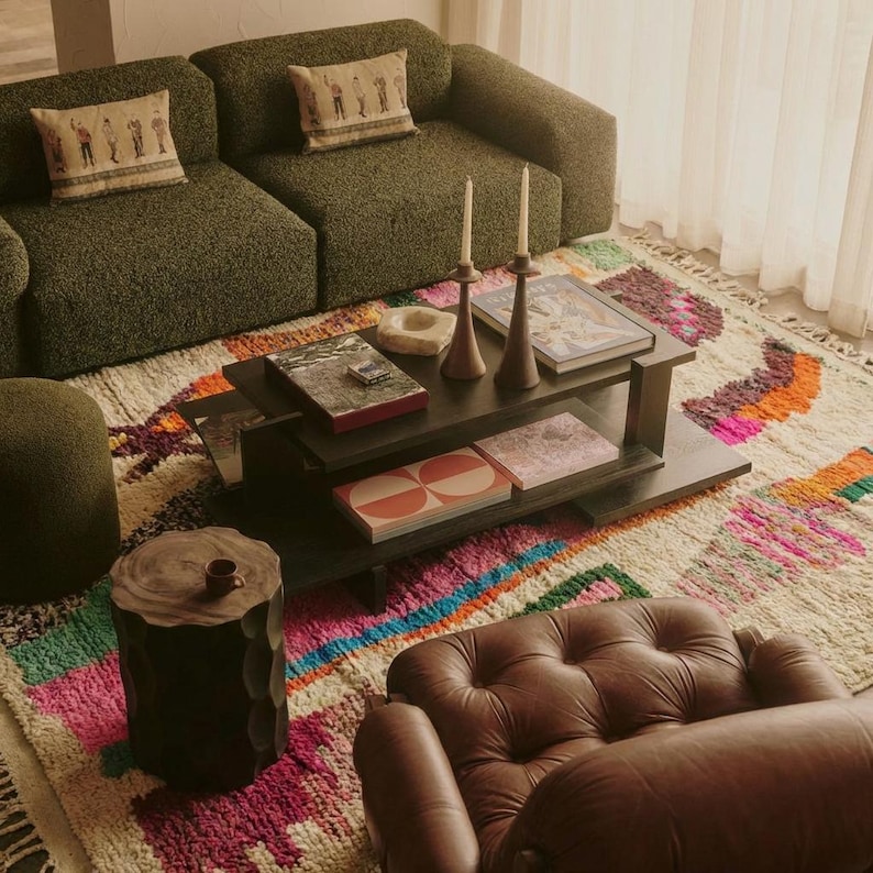A vibrant, high-pile handmade Moroccan wool rug displayed in a bright industrial studio against a white brick wall. The rug features an eclectic, abstract tribal design with bold splashes of neon pink, teal green, orange, royal blue, and charcoal grey on a natural cream base. The patterns include geometric shapes, abstract figures, and spontaneous color blocking. The rug showcases a thick, plush shaggy texture and authentic hand-knotted cream tassels at the bottom.