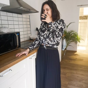 Peut inclure: Une femme porte un chemisier à imprimé floral noir et blanc et une longue jupe plissée bleu marine. Elle tient une tasse sombre dans une cuisine avec des armoires blanches et un comptoir en bois. Un micro-ondes et une plante sont également visibles.