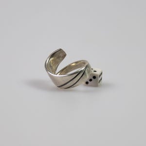 Könnte beinhalten: Ein silberfarbener Wickelring mit einem glatten, geschwungenen Band, das zwei dünne, dunkle eingravierte Linien auf der Oberfläche aufweist. Der Ring liegt neben einem kleinen, weißen sechsseitigen Würfel mit schwarzen Punkten, der drei Punkte zeigt.