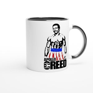 Può includere: Tazza in ceramica bianca con bordo e manico neri. La tazza presenta un'illustrazione in bianco e nero di un pugile che indossa pantaloncini da boxe rosso, bianco e blu. Il testo "APOLLO CREED" è stampato in nero sotto l'illustrazione.