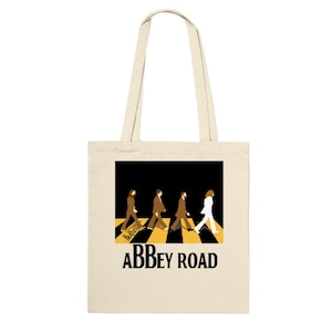 Può includere: Una borsa tote beige con un'immagine in bianco e nero dei Beatles che camminano su un attraversamento pedonale. Il testo "Abbey Road" è stampato in nero sotto l'immagine.
