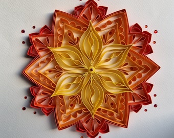 Puede incluir: Un mandala de flores de papel de varias capas en tonos naranja, amarillo y rojo. La flor está hecha de intrincados recortes de papel y es un hermoso ejemplo de arte en papel.