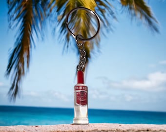 May include: A Smirnoff vodka bottle keychain with a red cap and a silver chain. The keychain is set against a backdrop of a tropical beach with palm trees and a blue sky.