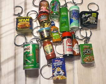 May include: A collection of miniature food and household product keychains featuring brands like Spam, Mentos, Breyers, Milo, Prego, Kraft, Lawry's, Tabasco, Apple Jacks, and Frosted Flakes. Each item is attached to a silver metal split ring keychain.