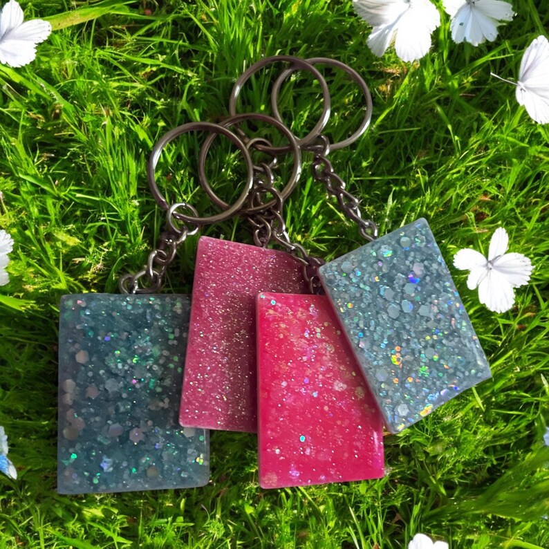 May include: Four rectangular keychains in shades of teal, pink, and red with glitter embedded in the resin. Each keychain is attached to a silver-colored metal ring. The keychains are displayed on a bed of green grass.