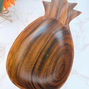 May include: A carved wooden bowl shaped like a pineapple, featuring a deep brown color with visible natural wood grain patterns. The bowl has a smooth, concave interior and a pointed top section that mimics the leaves of a pineapple fruit.