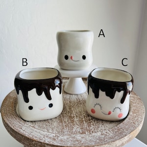 May include: Three ceramic marshmallow mugs with cute faces. The mugs are white with brown chocolate-like drippings. One mug has a pink tongue sticking out. The mugs are on a wooden tiered stand.