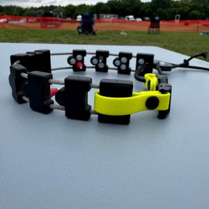 Puede incluir: Un dispositivo negro y amarillo con forma de collar con múltiples componentes rectangulares negros conectados por un cable plateado. El dispositivo tiene detalles en rojo y negro y un cierre amarillo. El fondo es una superficie gris con una escena exterior borrosa.