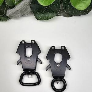 Op de afbeelding: Twee zwarte metalen clips met een draaibare lus aan de onderkant. De clips hebben een veerbelaste mechanisme en een vergrendelingsfunctie.