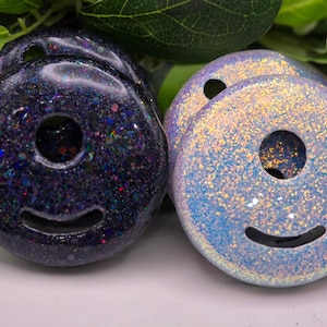 May include: Close-up of two glittery, donut-shaped objects with a smiley face design. One is dark blue with colorful glitter, the other is light blue with gold glitter. They have a hole in the center.