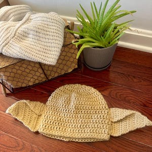 May include: A light tan crochet beanie featuring two large, pointed, triangular ear-like flaps extending from the sides. The hat is crafted with a textured stitch pattern and is positioned on a dark wood surface near a wire basket and a green plant.