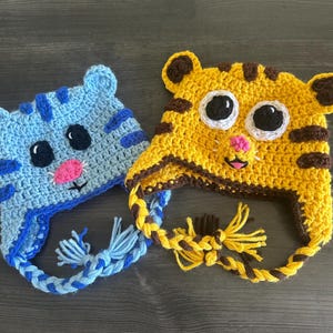 Puede incluir: Dos gorros de ganchillo hechos a mano. Uno es azul con rayas azul oscuro y una nariz rosa, el otro es amarillo con rayas marrones y una nariz rosa. Ambos tienen ojos negros grandes y orejeras con lazos trenzados.