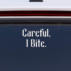 May include: White vinyl decal with the text "Careful, I Bite" in a gothic font, applied to the rear window of a white vehicle. The decal is centered and clearly visible against the dark glass.