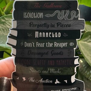 K&ouml;nnte beinhalten: Ein Aufkleber mit einem Stapel von B&uuml;chern mit Titeln wie "The Gallows" und "Isolation". Die B&uuml;cher sind &uuml;berwiegend dunkel gef&auml;rbt mit wei&szlig;em Text und Illustrationen, darunter eine Schlange. Das untere Buch tr&auml;gt die Aufschrift "Love in a Time of the Zombie Apocalypse."