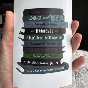 May include: A sticker depicting a stack of books with titles including "The Gallows" and "Isolation." The books are in shades of green, grey, and black, with white text. The sticker is held in a hand.
