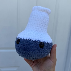 May include: A crochet plush toy shaped like a laboratory flask with a rounded bottom and a narrow neck. The bottom section is dark blue, and the top section is white. It features two small, round, black plastic safety eyes on the front.