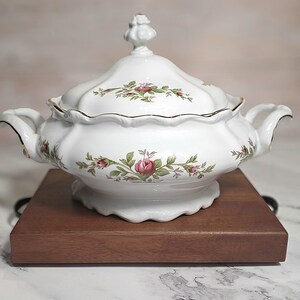 May include: White porcelain soup tureen with a lid, decorated with a floral rose pattern in pink and green. The tureen has gold trim and handles, on a dark wooden base. A classic piece for serving soups and stews.
