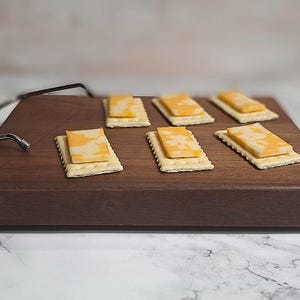 May include: A wooden serving board with six crackers topped with slices of yellow and white marbled cheese. The board has a dark brown finish and a metal handle. The crackers are arranged in two rows of three.
