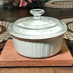 Puede incluir: Una cacerola de cerámica blanca con tapa de vidrio se encuentra sobre una base de madera. La base es de color marrón rojizo y está hecha de madera.