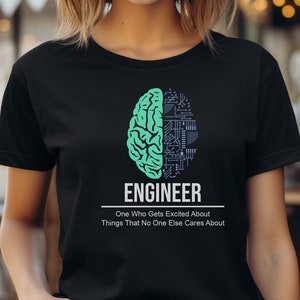 Può includere: Una maglietta nera con una grafica di cervello verde e nero e il testo "Ingegnere. Uno che si entusiasma per cose che a nessun altro importa."