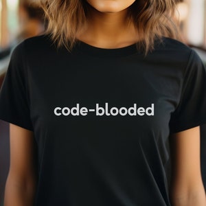 Op de afbeelding: Een vrouw draagt een zwart T-shirt met de tekst "code-blooded" in wit op de voorkant. Ze staat in een treinwagon met ramen aan beide kanten.