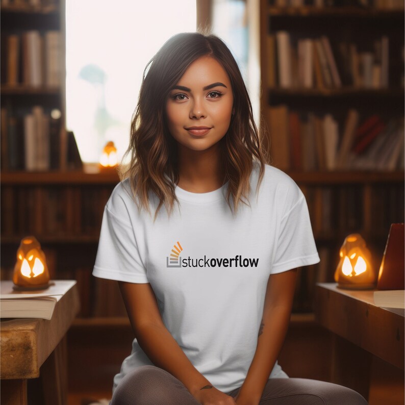May include: A woman wearing a white t-shirt with the Stack Overflow logo printed on the front. The logo is orange and black and features a stylised image of a book with a rising sun behind it. The text "stackoverflow" is printed below the image.