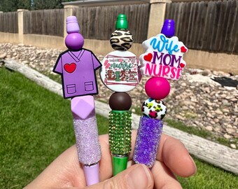 May include: Three pens featuring decorative silicone beads and charms. One pen has a purple scrub top charm, one has a round 'Nurse Life' charm with leopard print, and one has a 'Wife Mom Nurse' charm with red hearts. Each pen includes glittery accents.