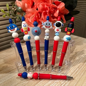 May include: Set of seven red and blue pens with different character toppers, including a shark, rocket, astronaut, and a baseball. The pens are displayed on a clear plastic stand.