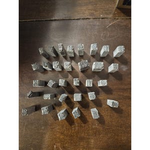 May include: A collection of thirty-one individual metal letterpress type blocks, each featuring a unique, ornate, and decorative serif letter embossed on the top surface. The blocks are rectangular, silver-toned, and arranged in a loose grid pattern.