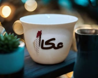 May include: A white ceramic coffee cup with Arabic text in brown and red. The cup is on a dark surface, with a small succulent plant in a blue pot to the left and a dark glass to the right. The background has blurred bokeh lights.