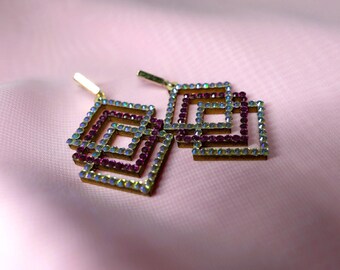 May include: A pair of gold-toned earrings with a geometric design. The earrings are made of multiple squares, each with a border of iridescent rhinestones and a center of pink rhinestones.