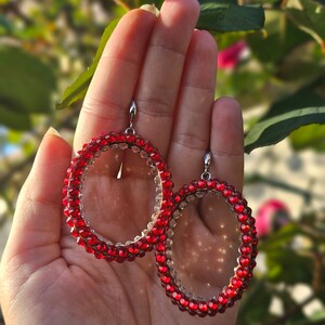 May include: A pair of red crystal hoop earrings with a clear center. The earrings are made of small, round crystals that are strung together to form a hoop shape. The earrings are held in a hand.