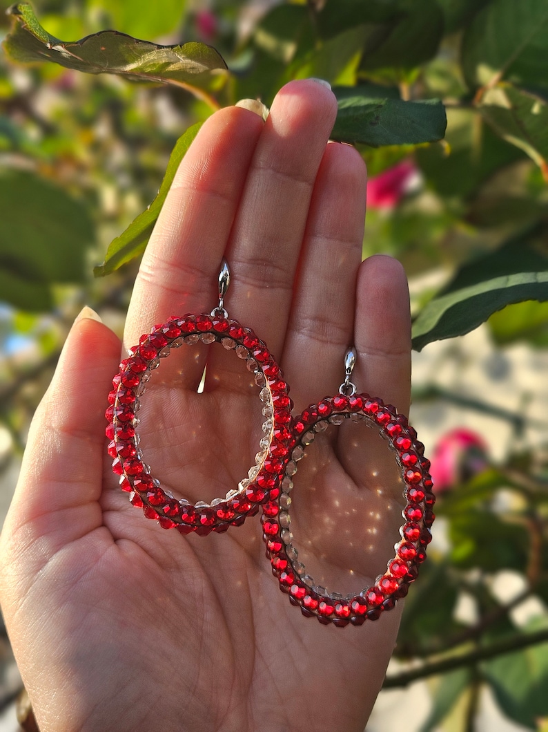 May include: A pair of red crystal hoop earrings with a clear center. The earrings are made of small, round crystals that are strung together to form a hoop shape. The earrings are held in a hand.
