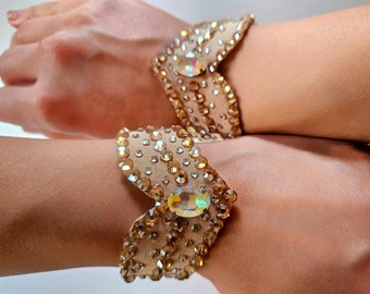 May include: A pair of beige fabric wrist cuffs adorned with sparkling rhinestones and iridescent crystals. The cuffs have a scalloped edge and a large, oval-shaped crystal in the center.