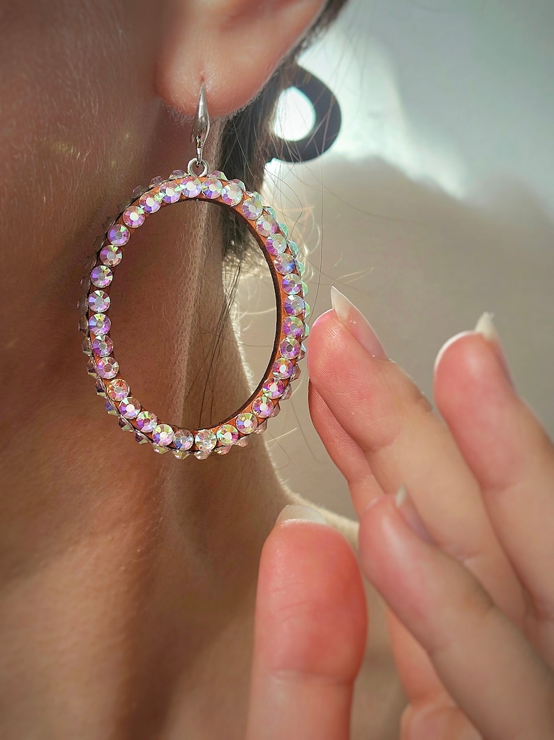 May include: A pair of large, oval-shaped earrings with a silver-toned metal setting. The earrings are adorned with iridescent rhinestones that create a sparkling effect.