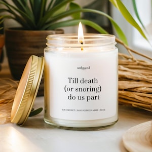 May include: A white soy and coconut candle in a clear glass jar with a gold lid. The candle has the text "Till death (or snoring) do us part" printed on the label. The candle is 2.5 ounces.