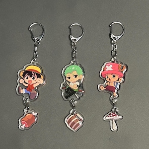 May include: Three acrylic keychains featuring anime characters. One keychain shows a character in a straw hat with a piece of meat. Another shows a character with green hair and a piece of meat. The third shows a character in a pink hat with a mushroom.
