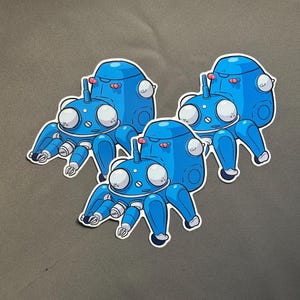 May include: Three blue robot stickers with white outlines are arranged on a gray surface. Each sticker features a rounded body, large white eyes, and multiple articulated legs. A small red antenna is on top of each robot.