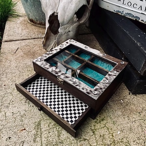 May include: A dark wooden jewelry box with a glass lid framed by cowhide and silver studs. The top section features a turquoise interior with grid dividers, while the bottom pull-out drawer is lined with a black and white checkered pattern.